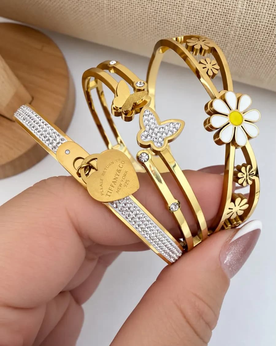 Braceletes dourados em aço, com detalhes de flor, borboleta e coração, segurados na mão